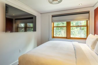 A bright bedroom in Cedar Crest Woodpecker Cottage 7 with a large window overlooking greenery, a wall-mounted TV, and a bed with white linens.