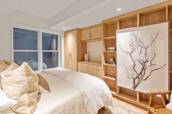 A cozy bedroom with a bed, built-in wooden cabinetry, a large window, and a nature-inspired wall art.