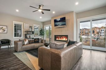 A luxurious and cozy living room with two brown leather sofas, a modern fireplace, a wall-mounted TV, and large windows looking out onto an urban setting.