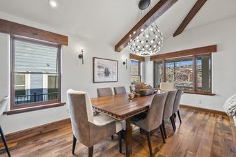 The spacious dining area at Park Avenue Home featuring a large wooden dining table with eight upholstered chairs, illuminated by a contemporary chandelier, and set against a backdrop of exposed wooden beams and a large window.