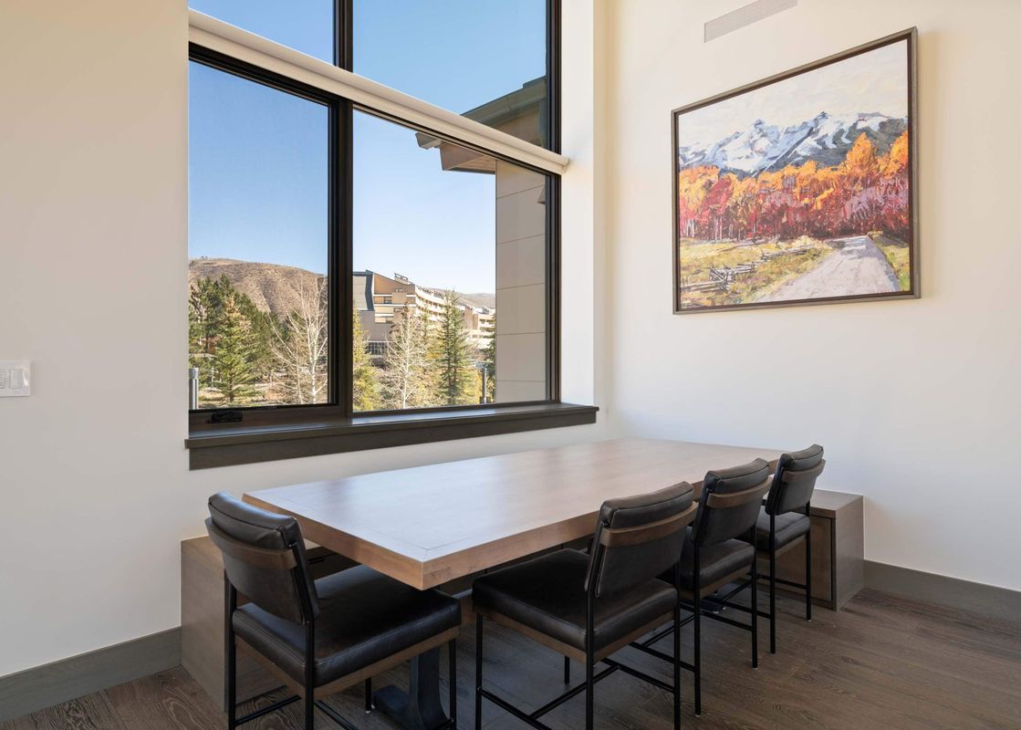 A dining area with a long wooden table and four black bar stools, featuring large windows with mountain views and a landscape painting on the wall.