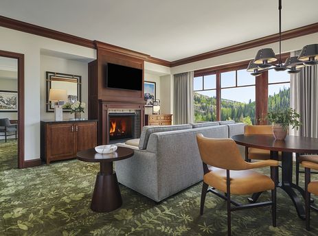 The living area of the Montage Deer Valley One Bedroom Suite Vista View features a comfortable sofa and armchairs, a large flat-screen TV above a lit fireplace, and a dining table with seating for four, all set on a vibrant green botanical carpet.