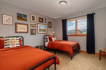 The image features a cozy bedroom at 2002 Lupine Lane, boasting two single beds with vibrant orange duvets and an assortment of framed artwork adorning the light gray walls. A window with dark blue curtains provides a glimpse of the surrounding neighborho