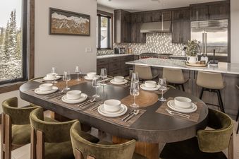 The spacious kitchen and dining area feature dark wood cabinetry, a large marble island, and a dining table set with white dinnerware and olive green chairs. The large window overlooks snow-covered trees.