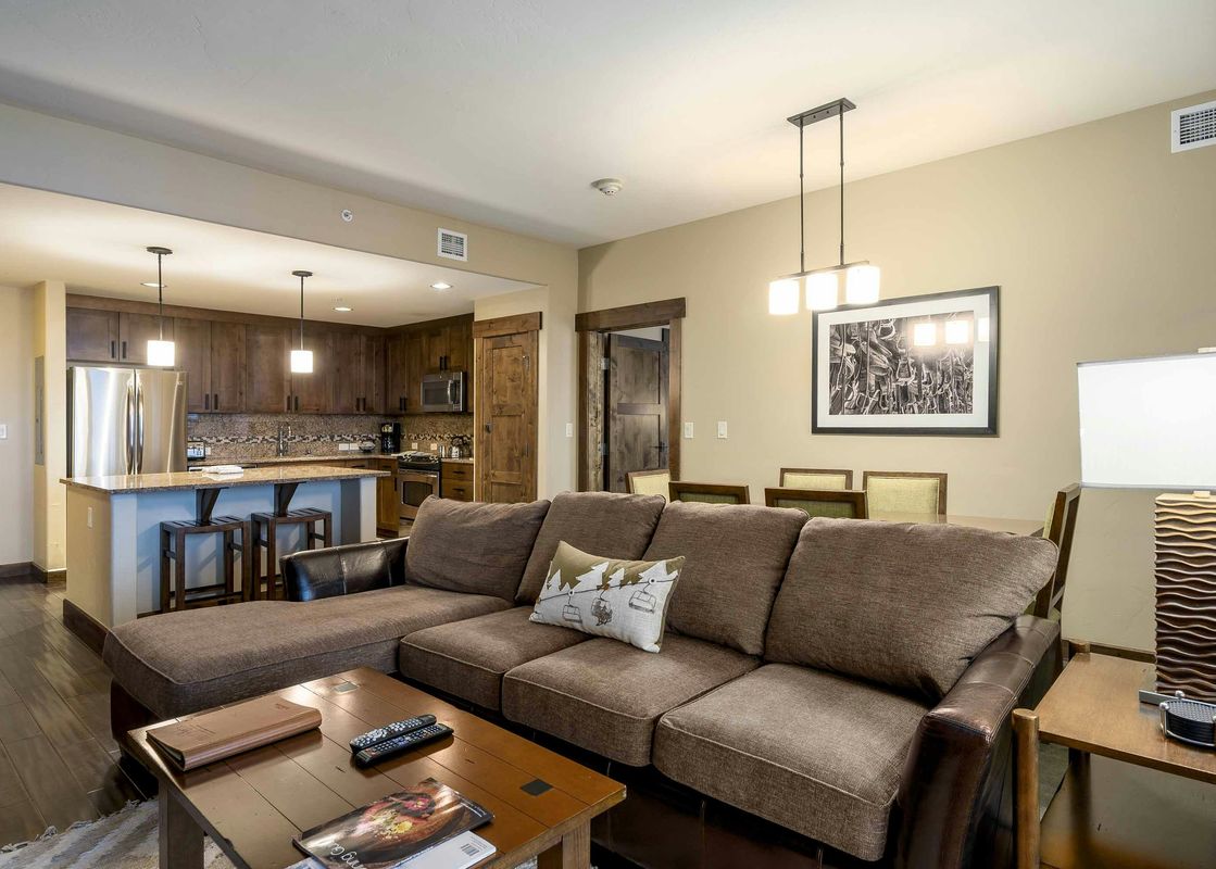 The Trailhead Lodge 4115 living room features a sectional sofa with a large pillow, a coffee table with books and remote controls, and a large window that overlooks the surrounding area.