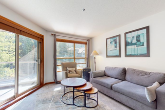 A spacious and inviting living area featuring a large grey sofa, a set of three round nesting coffee tables, a comfortable armchair, and large windows offering a view of lush greenery.