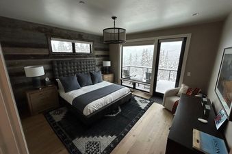 The master bedroom at Moonlight Basin 7 Deep Forest has a plush king-sized bed, a fireplace, a large window with a view of the snowy mountains, a wood-paneled accent wall, and a dresser with a lamp.