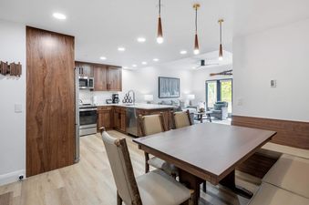 An expansive open-concept living area at The Charter E1305, featuring a modern kitchen with wooden cabinets, a dining area with a rustic table and chairs, and a cozy lounge with a sofa and armchair, all bathed in natural light from the glass door.