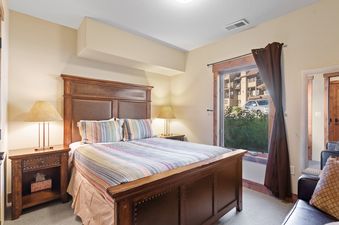A king bedroom at Pines Unit 240 features a wooden bed with a striped duvet, a leather headboard, and a nightstand with a lamp, with a window offering an exterior view.
