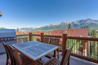 The outdoor dining area on the balcony of Tristant 125 features a wooden table and chairs, with a barbecue grill visible to the left, all set against a stunning mountain backdrop.