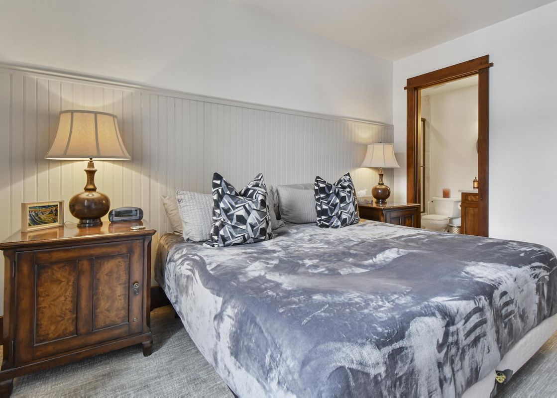 A king bedroom in Arrowleaf Lodge 311 with a grey and white bed, flanked by wooden nightstands and lamps, with a doorway leading to the bathroom.