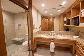 The spacious bathroom features a double vanity with granite countertops, a large mirror, a separate toilet, and a glass-enclosed walk-in shower with a built-in bench.