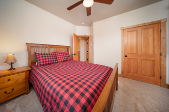 A cozy bedroom featuring a wooden bed frame with a headboard and footboard, dressed in a vibrant red and black buffalo check duvet and matching pillows, and complemented by a rustic wooden nightstand with a lamp.