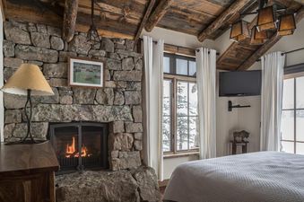 A cozy bedroom with a stone fireplace, a large bed with a quilted spread, and windows showcasing a snowy winter landscape.