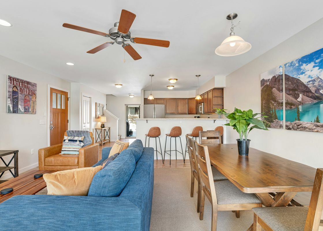 An inviting open concept living area features a blue sofa, a wooden dining table, and a well-equipped kitchen, all bathed in warm lighting.