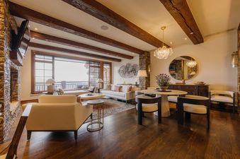 A wide shot of the living room and dining area, featuring wooden beams, a stone fireplace, large windows with mountain views, and modern furnishings.