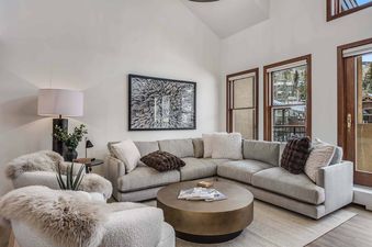 A cozy and elegant living room in Woodrun Place 56 with a large sectional sofa, a round coffee table, and armchairs, featuring large windows overlooking a snowy landscape.