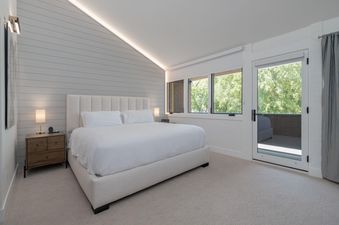 A modern bedroom with a king-size bed, white shiplap accent wall, subtle cove lighting, and large windows looking out onto green trees.