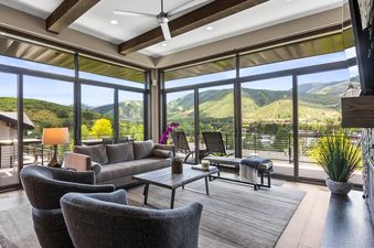 A spacious and modern living room with floor-to-ceiling windows offering panoramic mountain views, featuring a grey sofa, two armchairs, a wooden coffee table, and a ceiling fan.