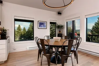 A modern dining room with a round wooden table, black metal chairs, and a window with a view of a forest and mountains.