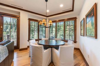 A dining room table and chairs are set in front of large windows with blinds.