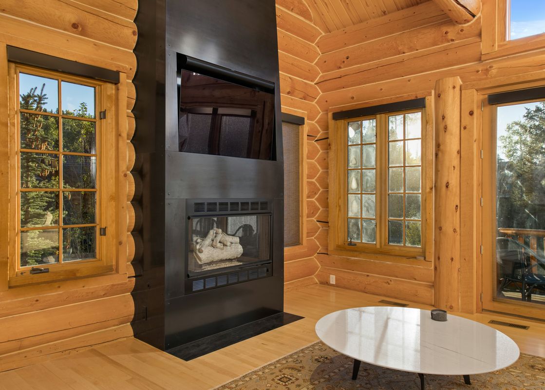 A cozy living area at Sundance Lodge 108 Lodges Lane with a modern black fireplace, log walls, large windows showcasing green outdoors, and a round white coffee table.