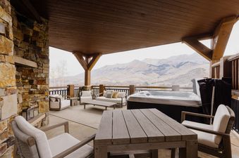An expansive outdoor patio with a hot tub, comfortable seating, a dining table, and a stone fireplace, all set against a stunning mountain backdrop.