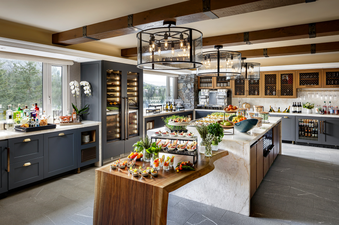 A well-lit, spacious kitchen and bar area featuring a long wooden and marble counter filled with various small appetizers, a bar setup with liquor bottles, and wine chillers, all with a view of snow-covered trees outside.