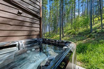 The hot tub at Wood Run Ski Haus, set against a wooden cabin wall, offers a relaxing soak amidst a lush green forest under a bright sky.