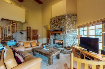 The spacious living area features a comfortable L-shaped leather sofa, a rustic wooden coffee table, a large stone fireplace, and a television, with a spiral staircase leading to an upper level.