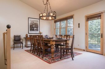 Warmly lit dining room in Moose Creek 33 with a rustic wooden table, an artistic chandelier, and a glass door overlooking green pine trees.