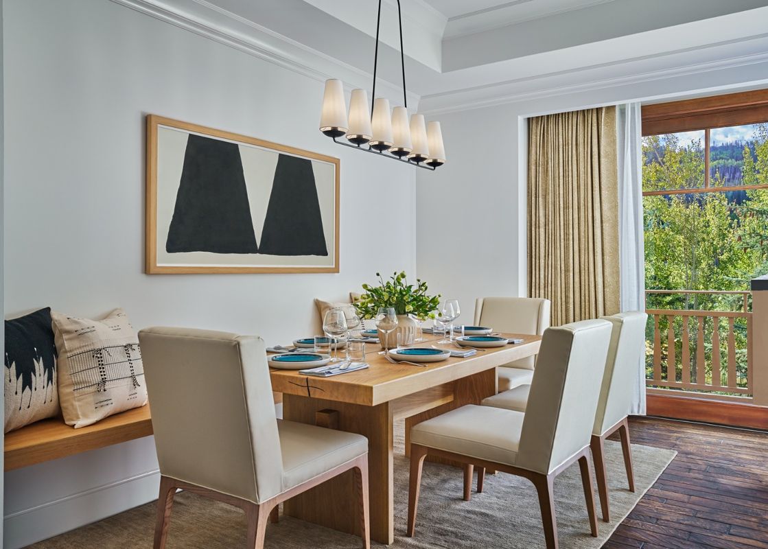 A modern dining area with a light wood table set with white and blue dishes, surrounded by cream-colored chairs and a built-in bench with decorative pillows.