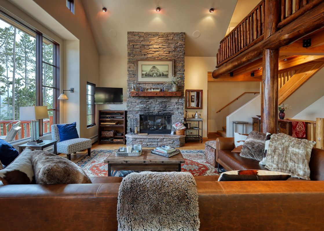 A spacious and cozy living room featuring a stone fireplace, leather sofas, and large windows overlooking a pine forest.