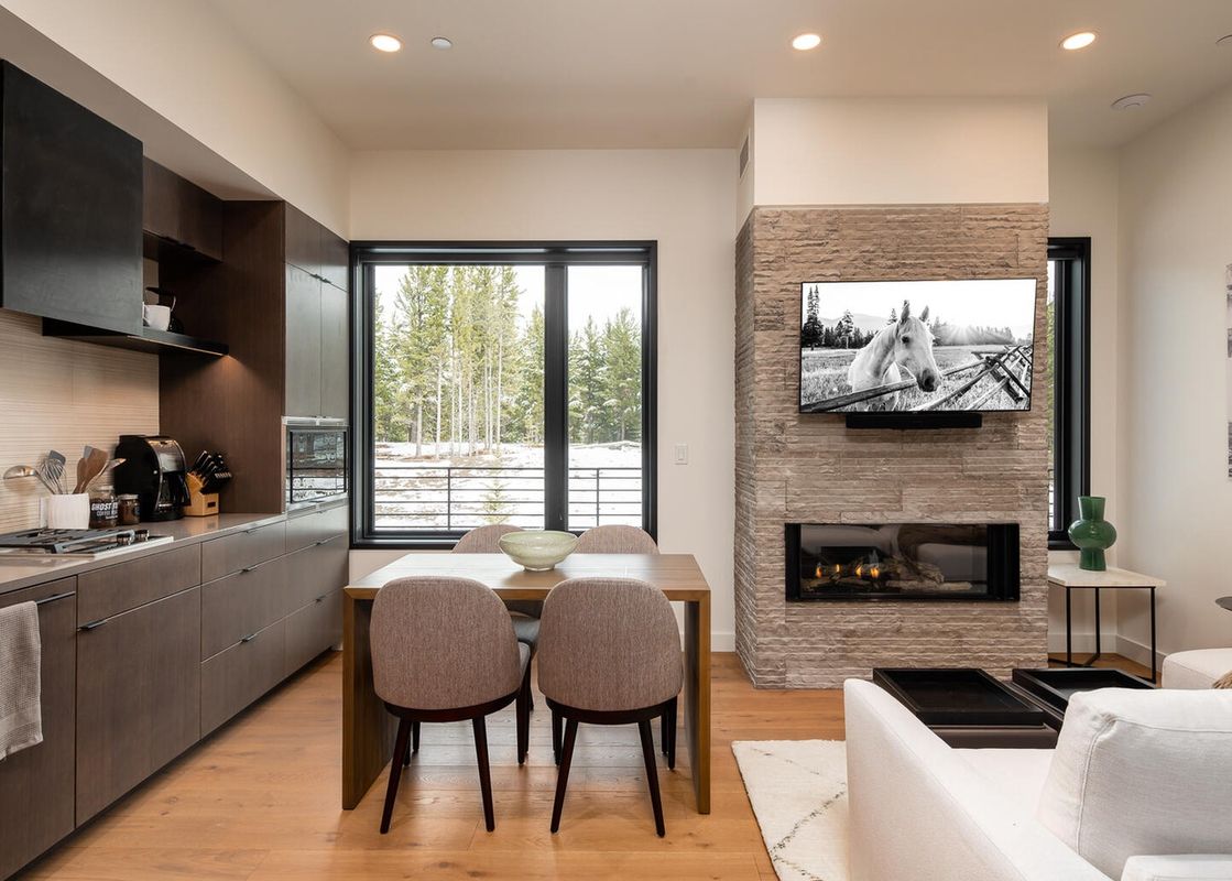 The kitchen and living area of 230 LakeLodge, showcasing modern design, a dining table, and a stone fireplace with a TV and a view of snow-dusted trees outside the window.
