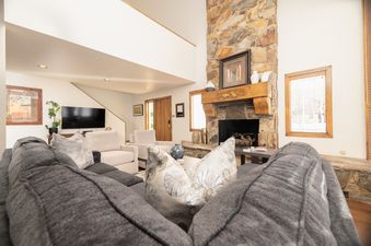 A spacious living room at Snow Park Chalet featuring a large stone fireplace, a gray sectional sofa with throw pillows, cream armchairs, and a flat-screen television.