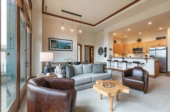 Living room with a grey sofa and brown leather armchair, a wooden coffee table, and a large window looking out onto a balcony.