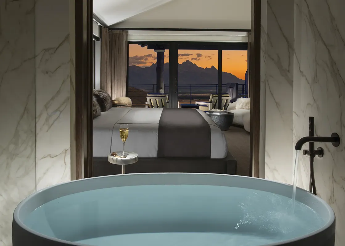 A luxurious bathroom with a freestanding tub filling with water, looking into a bedroom with a king-sized bed and a balcony with mountain views at sunset.