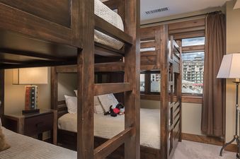 A cozy bunk room at Longs Peak One Steamboat Place 609 features dark wood bunk beds, warm lighting from two lamps, and a window offering a view of the surrounding buildings.