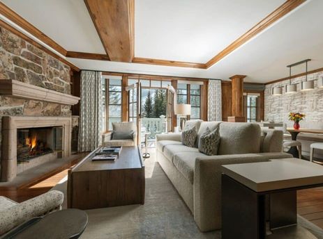 A cozy and elegant living room in Chateau Beaver Creek Residence 204, featuring a stone fireplace, comfortable seating, and large windows with a view of snow-covered trees.