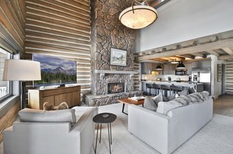 A spacious, open-concept living room with a large stone fireplace, two comfortable sofas, a TV, and an adjoining modern kitchen, all set against a backdrop of rustic log walls.