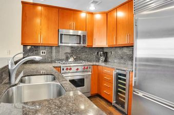The kitchen at Lowell 304 features a double basin stainless steel sink, a modern faucet, wood cabinets, granite countertops, stainless steel appliances, and a built-in wine cooler.