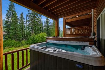 The private hot tub on the outdoor deck at Silvertip Ski Haus is partially covered and ready for a soothing soak, surrounded by a lush green landscape and towering evergreen trees.