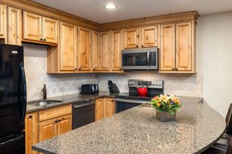 A modern kitchen with natural wood cabinets, granite countertops, and stainless steel appliances, including a refrigerator, microwave, oven, and dishwasher. A red kettle sits on the stovetop, and a floral arrangement adorns the breakfast bar.
