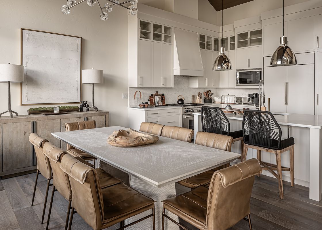 The open concept kitchen and dining area in Inspiration Point Townhome 4C features a long, light wood dining table with eight tan leather chairs, complemented by two black woven bar stools at the kitchen island, all under the glow of modern chandeliers an