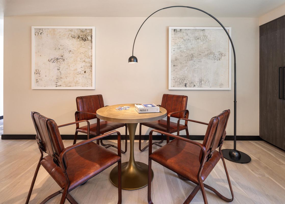 A game table with four brown leather chairs and a puzzle box, illuminated by a modern black arched floor lamp, set against light walls with abstract art.