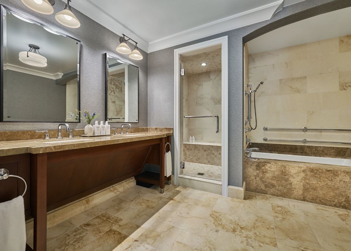 A luxurious bathroom with a double vanity, a walk-in shower, and a bathtub with grab bars, all in a neutral color palette with marble finishes.