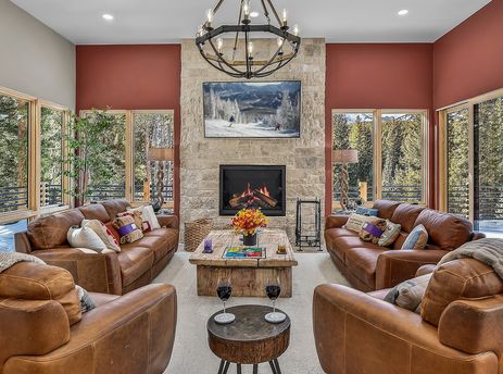 A grand living room in the Everwild Chalet, featuring brown leather furniture, a stone fireplace, and large windows looking out onto a snowy pine forest.
