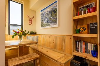 A cozy dining nook at Basecamp Luxe features built-in wooden benches around a rustic wooden table, with a large window and a bookshelf stocked with board games.