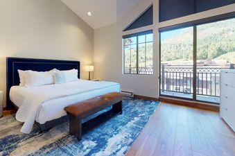 A luxurious master bedroom in Landmark 704 featuring a king-sized bed with a blue velvet headboard, an elegant nightstand, a brown leather bench, and large windows offering stunning mountain views.