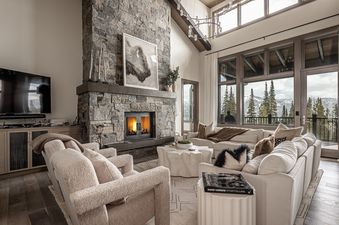 A spacious living room at Inspiration Point Townhome 4C features a stone fireplace, cream-colored seating, and large windows offering panoramic mountain and forest views.